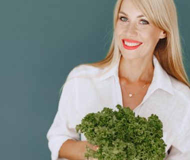 Cheerful woman with blonde hair holding fresh lettuce, promoting healthy living in a studio setting.