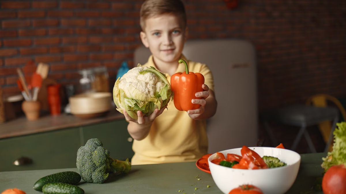 Little child demonstrating fresh red pepper and cauliflower and looking at camera while standing near table with vegetables and ingredients put into deep ceramic salad bowl in kitchen